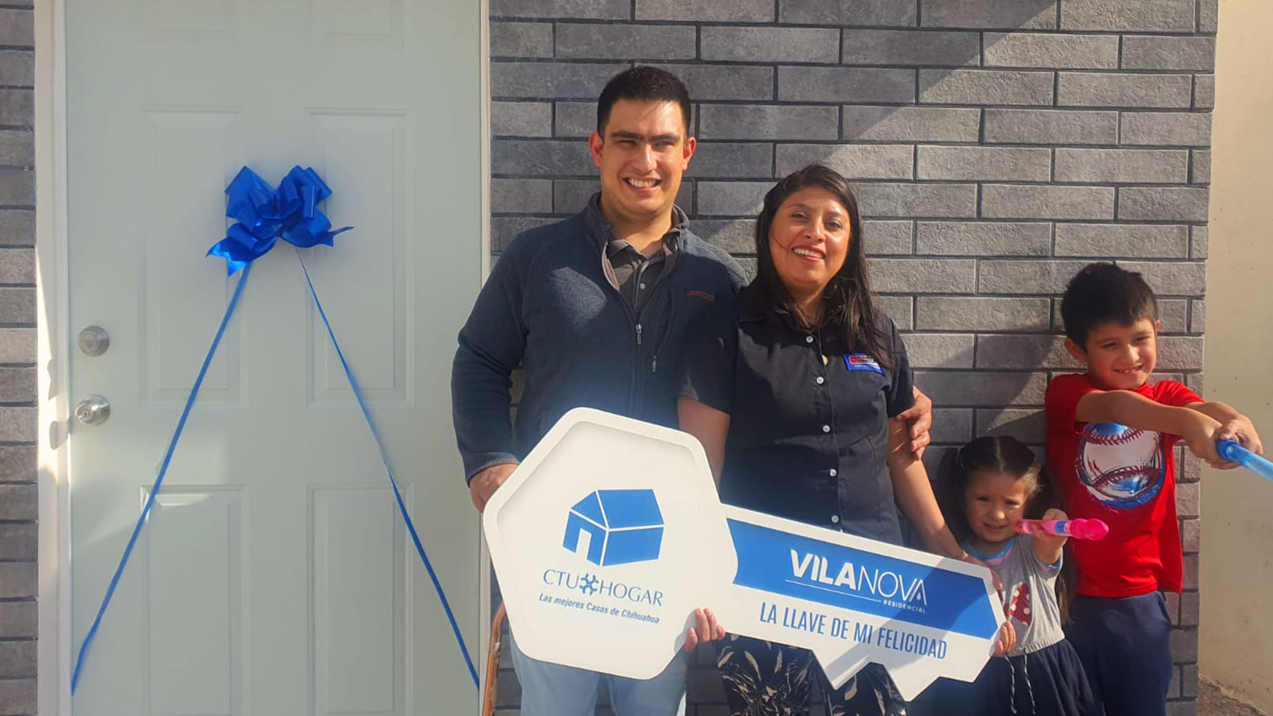 Familia de papá, mamá, niño de 8 años y niño de 4 años están felices de que les estén entregando la llave de su casa, frente a la puerta de entrada de su nueva propiedad.