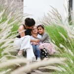 Madre abraza a sus dos hijos menores de 7 y 6 años en el patio de su casa, con varias plantas rodeándolos.