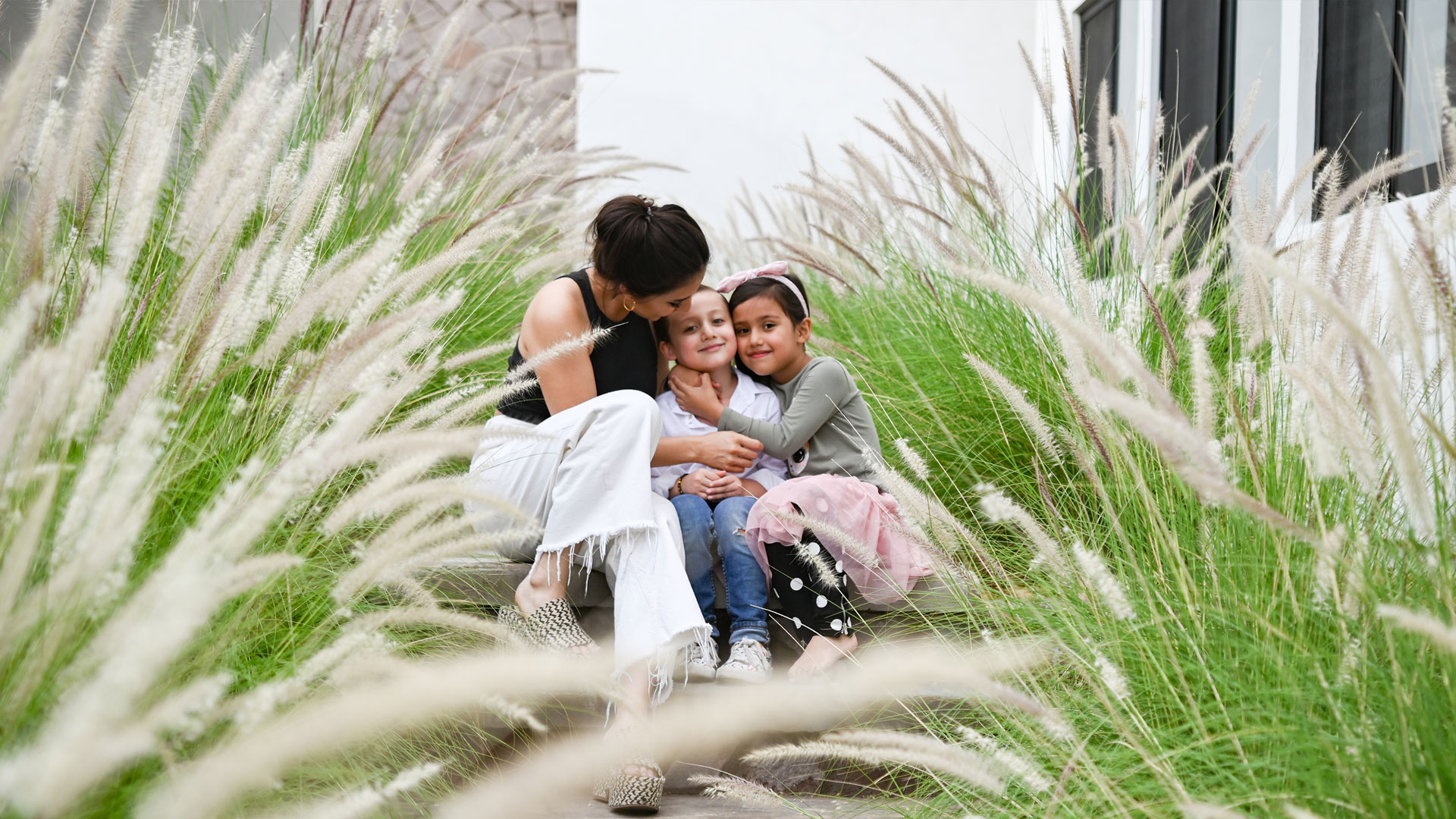 Madre abraza a sus dos hijos menores de 7 y 6 años en el patio de su casa, con varias plantas rodeándolos.