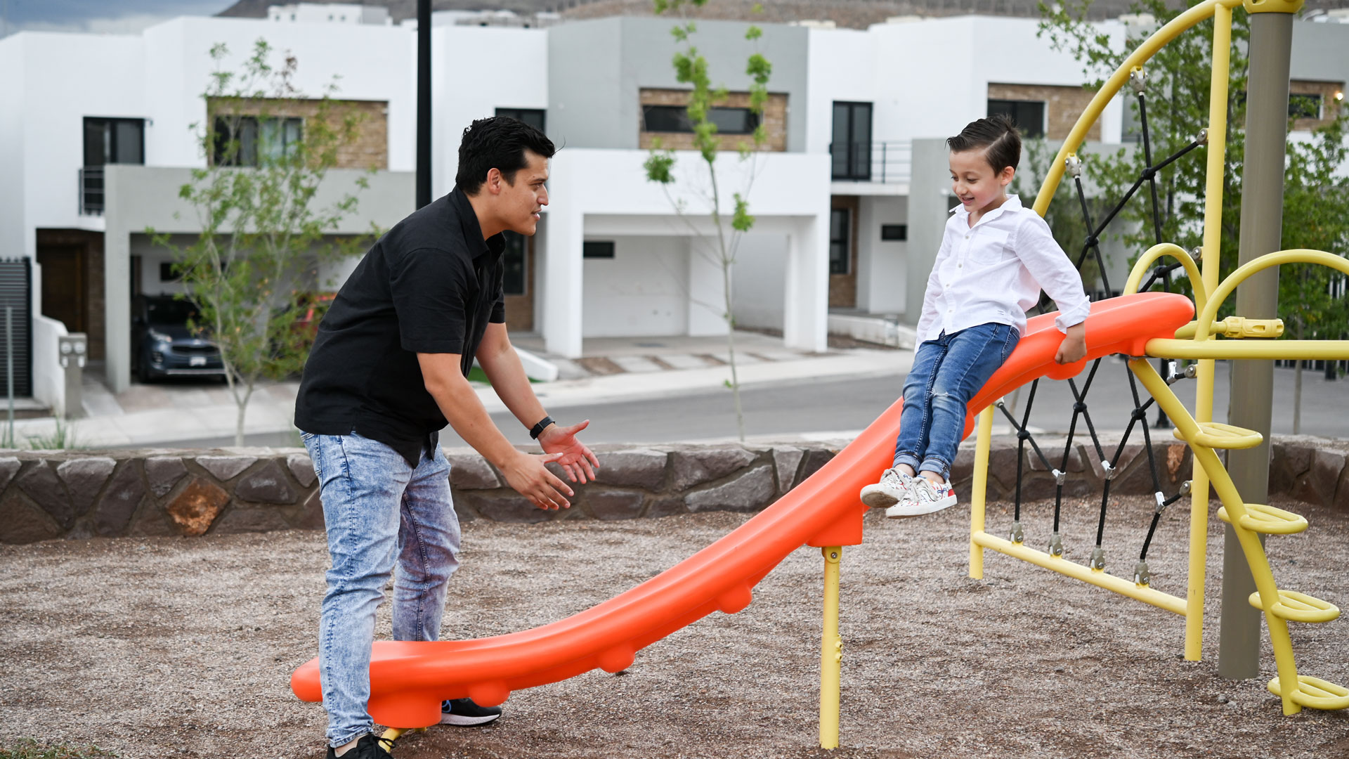 Un padre de familia de cerca de 30 años juega con su hijo de 6 años en un parque mientras su hijo se desliza en un rebaladero.